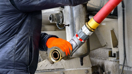 Une personne portant des gants orange connecte un tuyau rouge à un réservoir métallique.