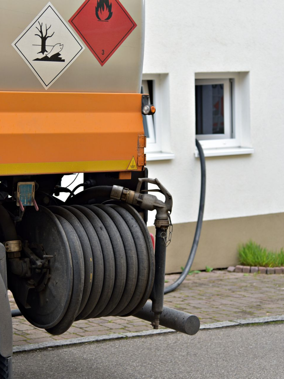 Camion-citerne avec tuyau ; bâtiment blanc en arrière-plan. Panneaux indiquant les risques d'inflammabilité et de pollution.