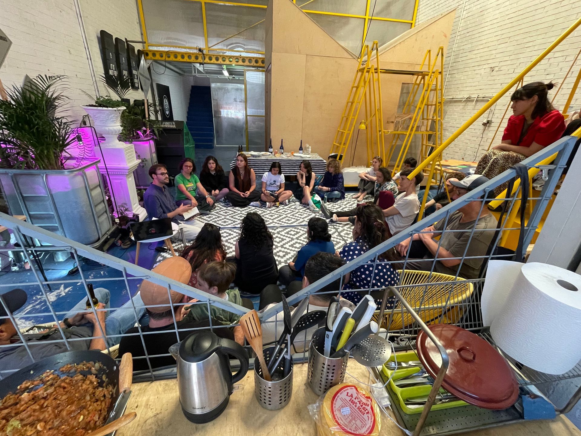 Grupo de personas sentadas en círculo en un interior. Comida y utensilios de cocina en primer plano.