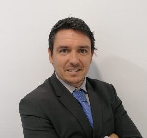 Retrato de un hombre con traje oscuro y corbata azul contra una pared blanca lisa.