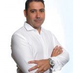 Hombre con camisa blanca y brazos cruzados, de pie sobre un fondo claro liso.