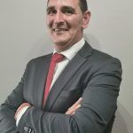 Hombre con traje oscuro y corbata roja, sonriendo con los brazos cruzados sobre un fondo claro liso.