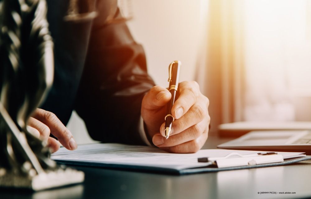 Eine Person im Anzug schreibt an einem Schreibtisch mit einem Stift auf Papier; im Hintergrund ist Sonnenlicht zu sehen.