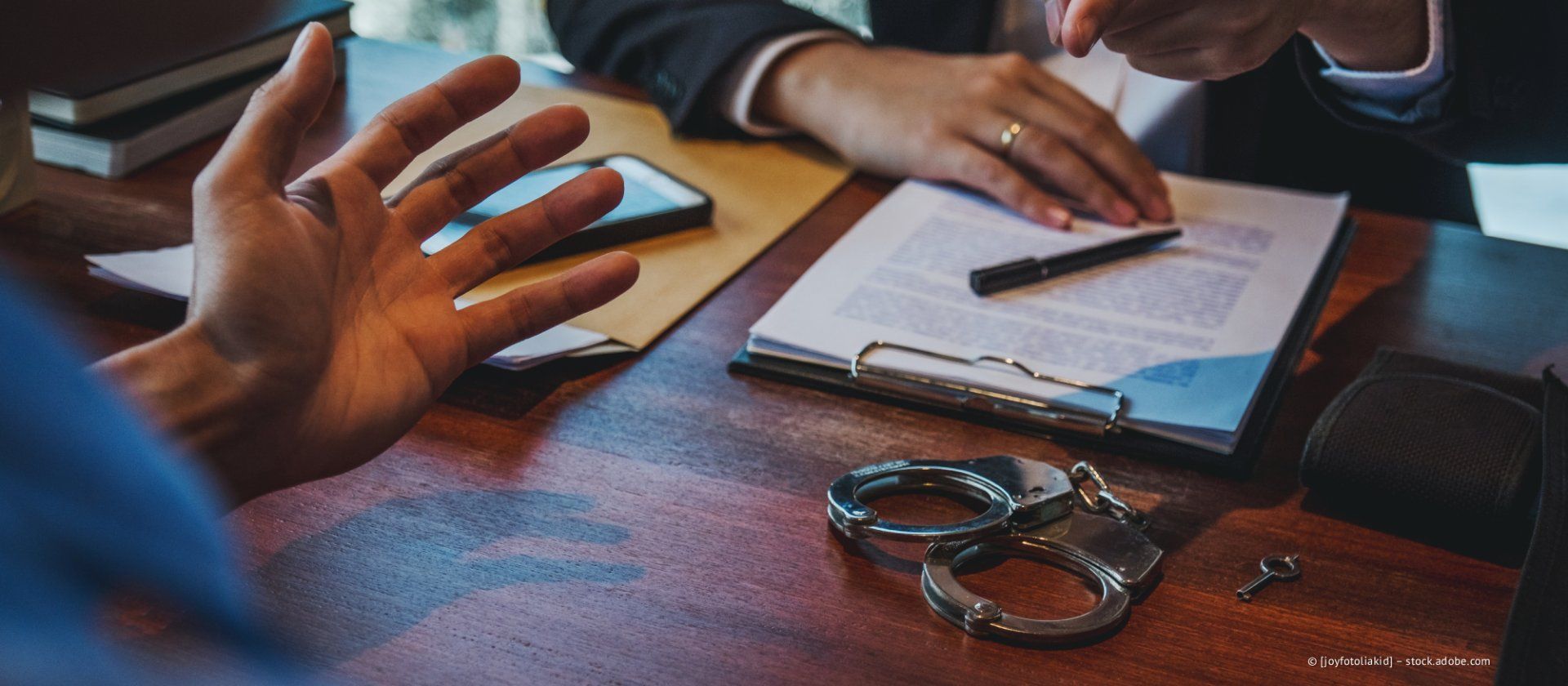 Nahaufnahme einer Hand, die über einen Tisch mit einem juristischen Dokument, einem Stift und Handschellen gestikuliert.