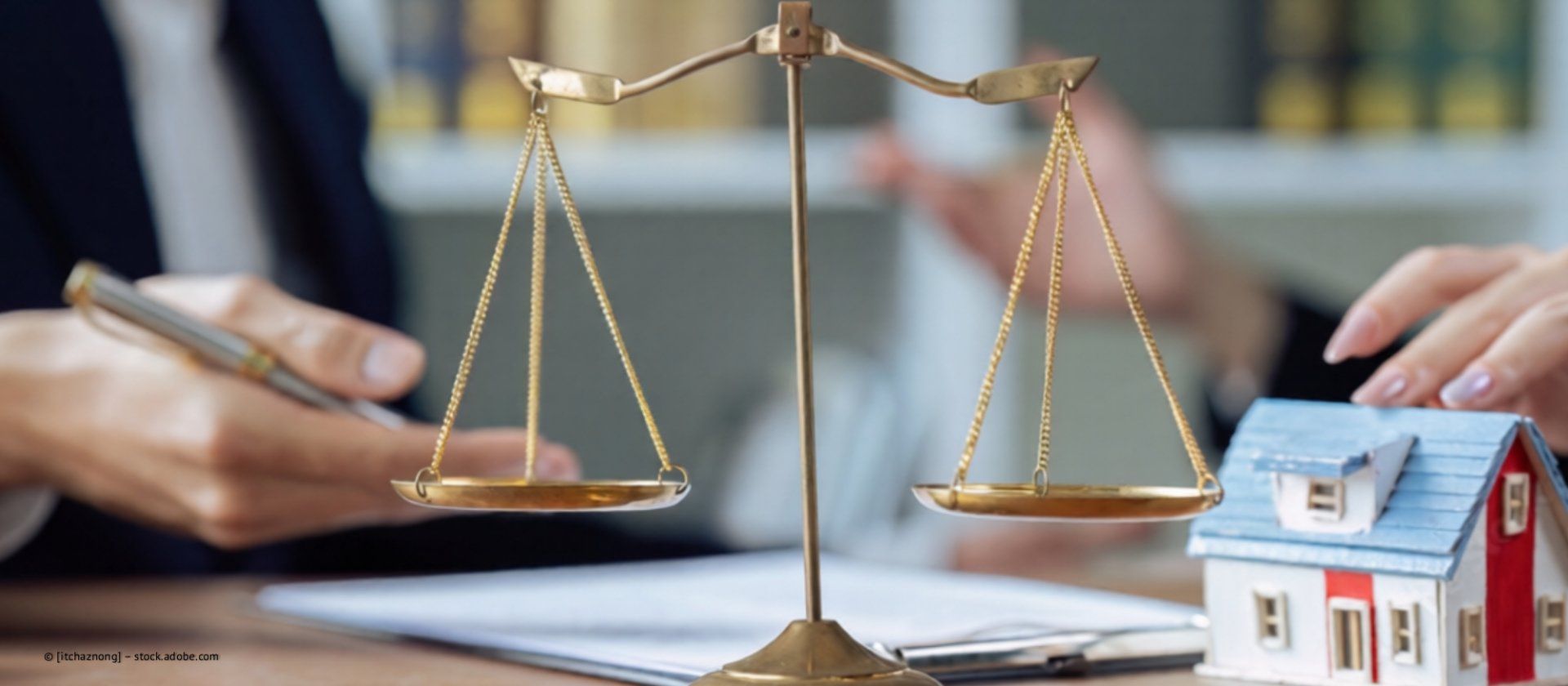 Eine Justitia-Waage vor einem Musterhaus, über die zwei Personen diskutieren.