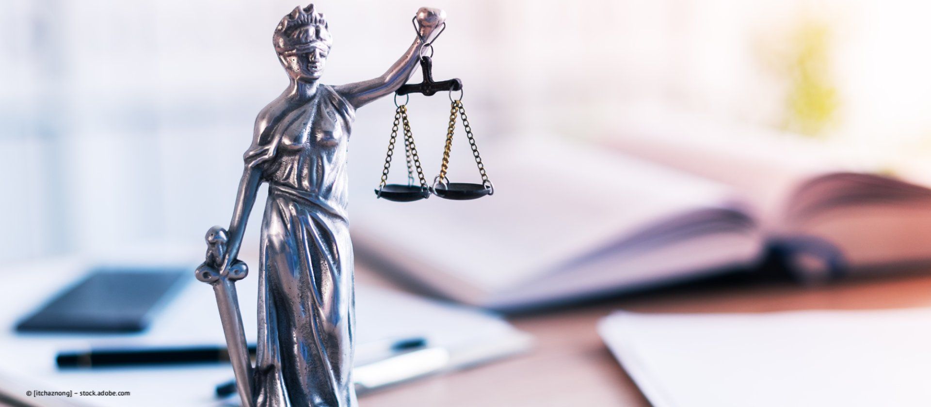 Statue der Justitia mit Waage und Schwert, im Hintergrund Bücher und Papiere.