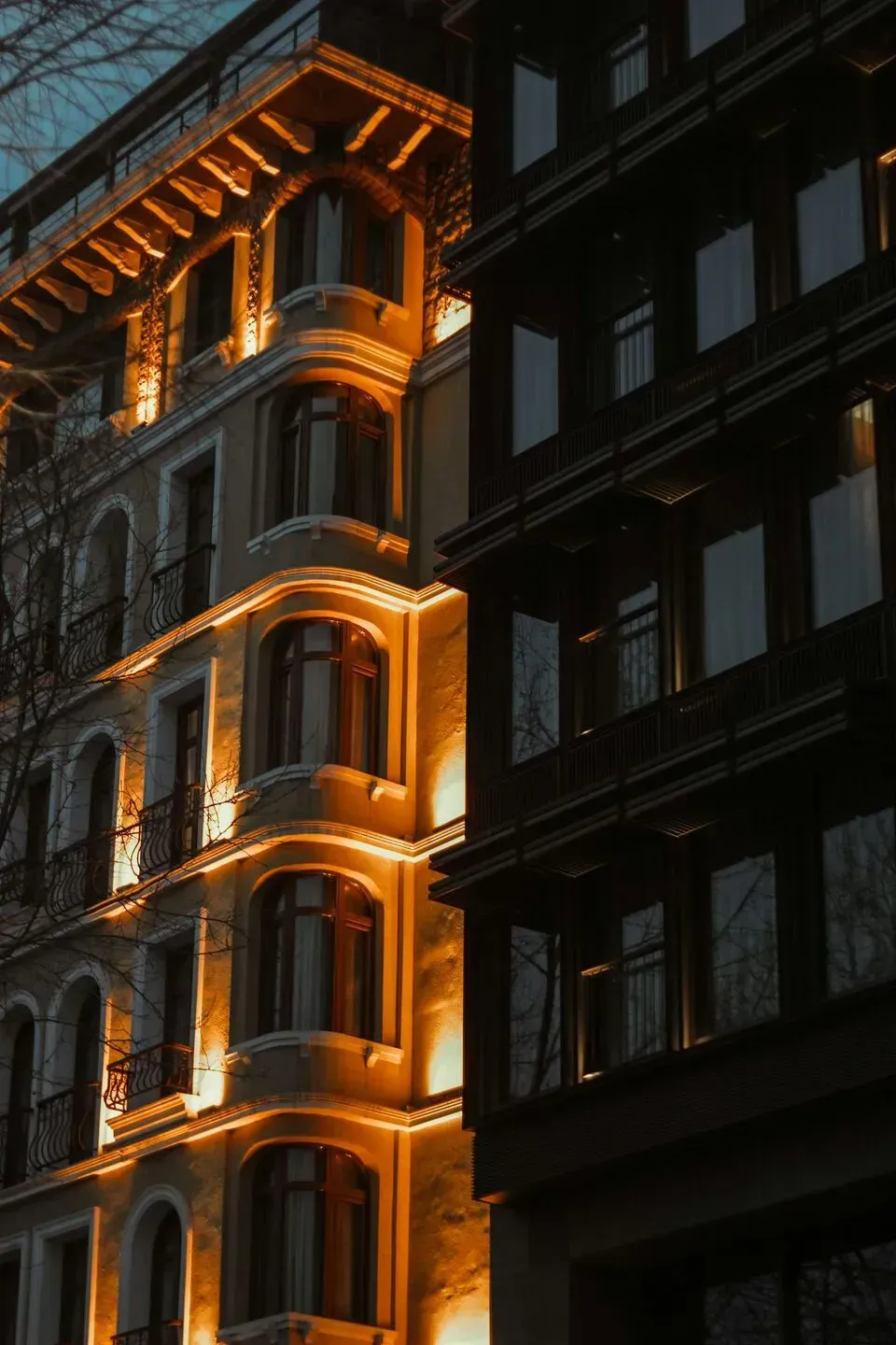 Un edificio de piedra ornamentado y bien iluminado por la noche se encuentra al lado de un edificio oscuro y moderno con ventanas de vidrio reflectante.