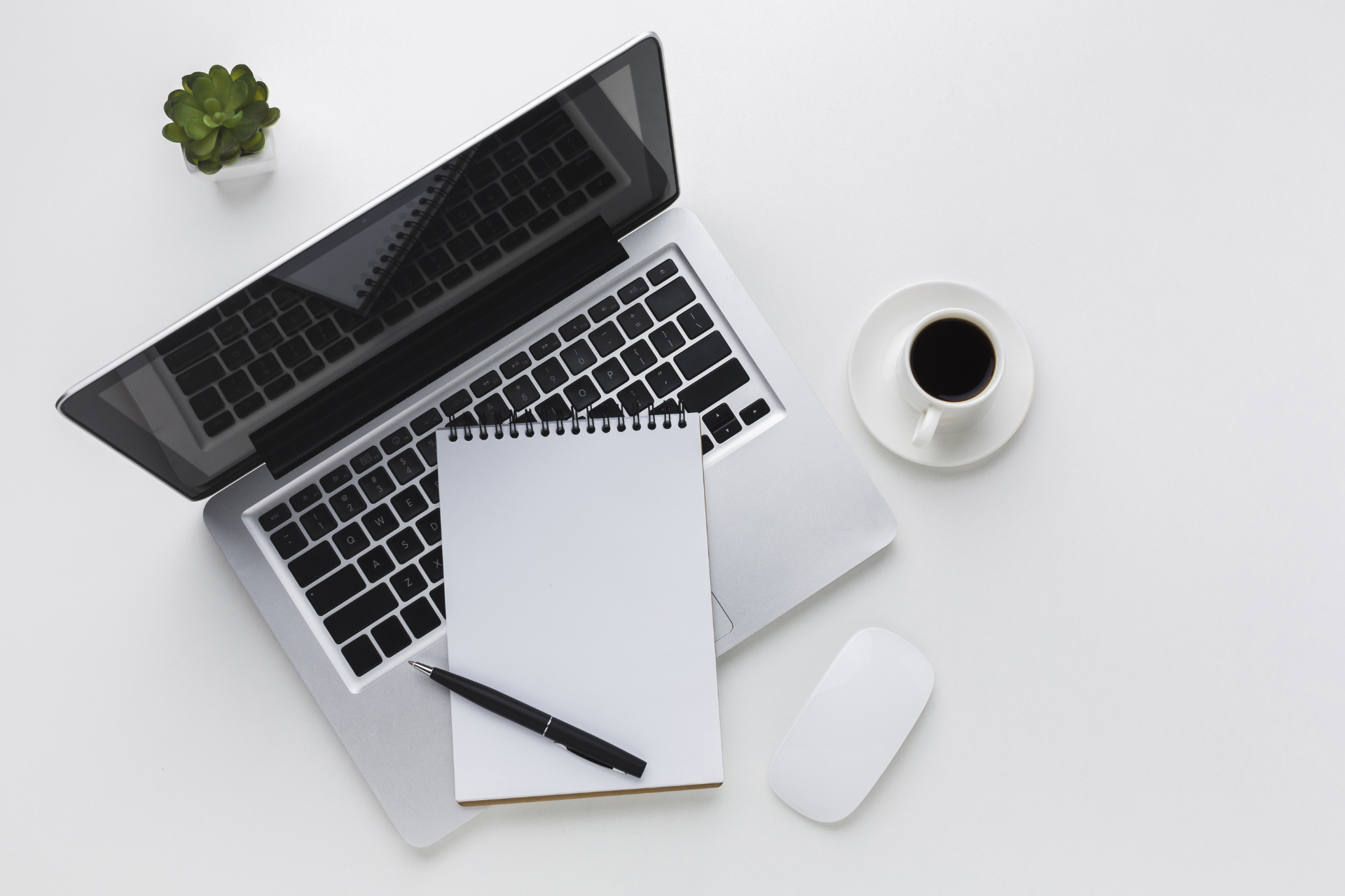 Una computadora portátil, un cuaderno, un bolígrafo, un ratón y una taza de café sobre una mesa blanca.