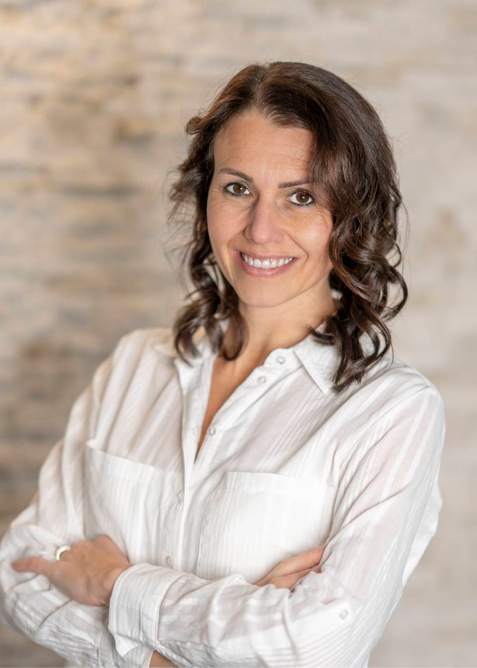 Frau mit braunen lockigen Haaren, die Arme verschränkt, lächelnd, trägt ein weisses Hemd mit Knöpfen.