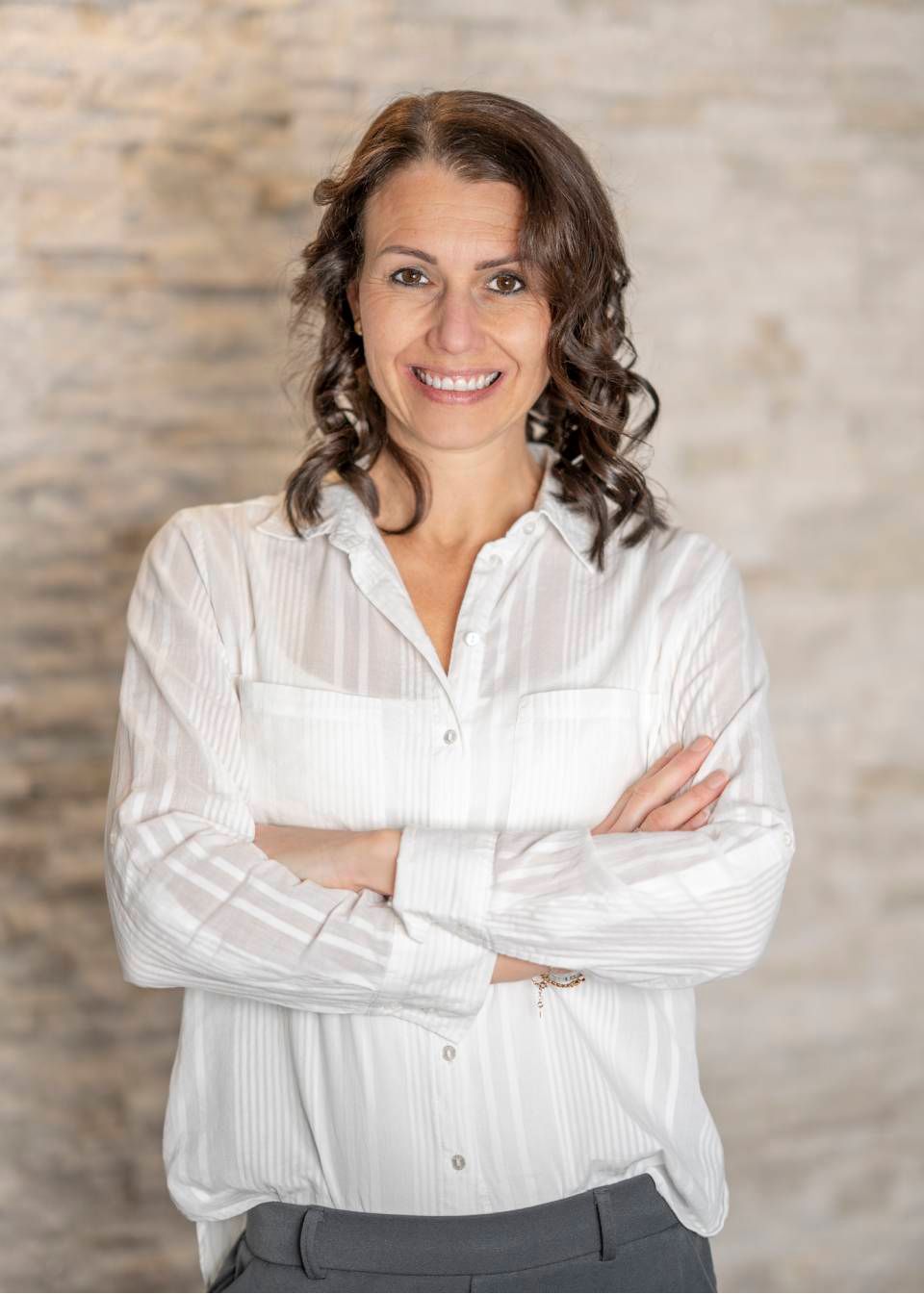 Frau mit verschränkten Armen, lächelnd, in einem weissen Hemd mit Knöpfen, steht vor einer hellen Wand.