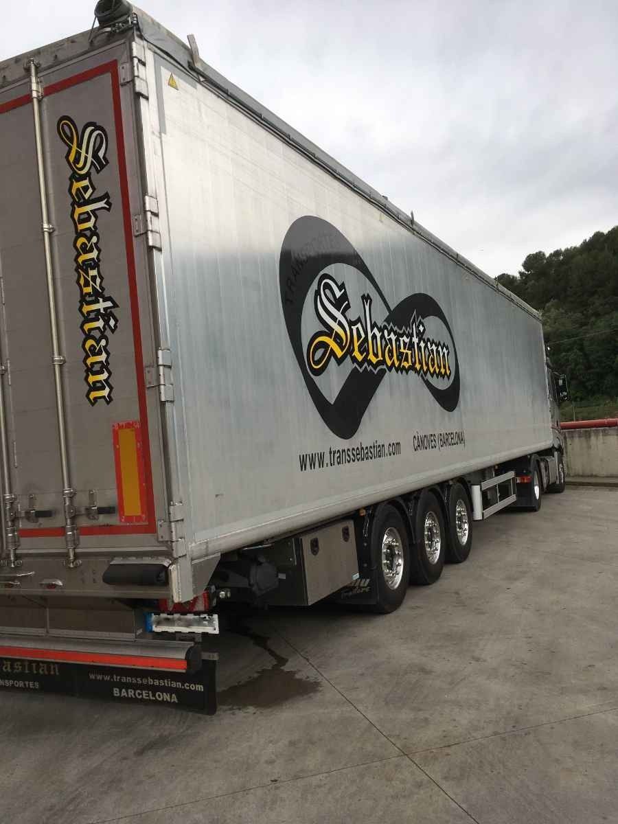 Semirremolque plateado con un gran logotipo negro y amarillo estacionado en un terreno pavimentado bajo un cielo nublado.