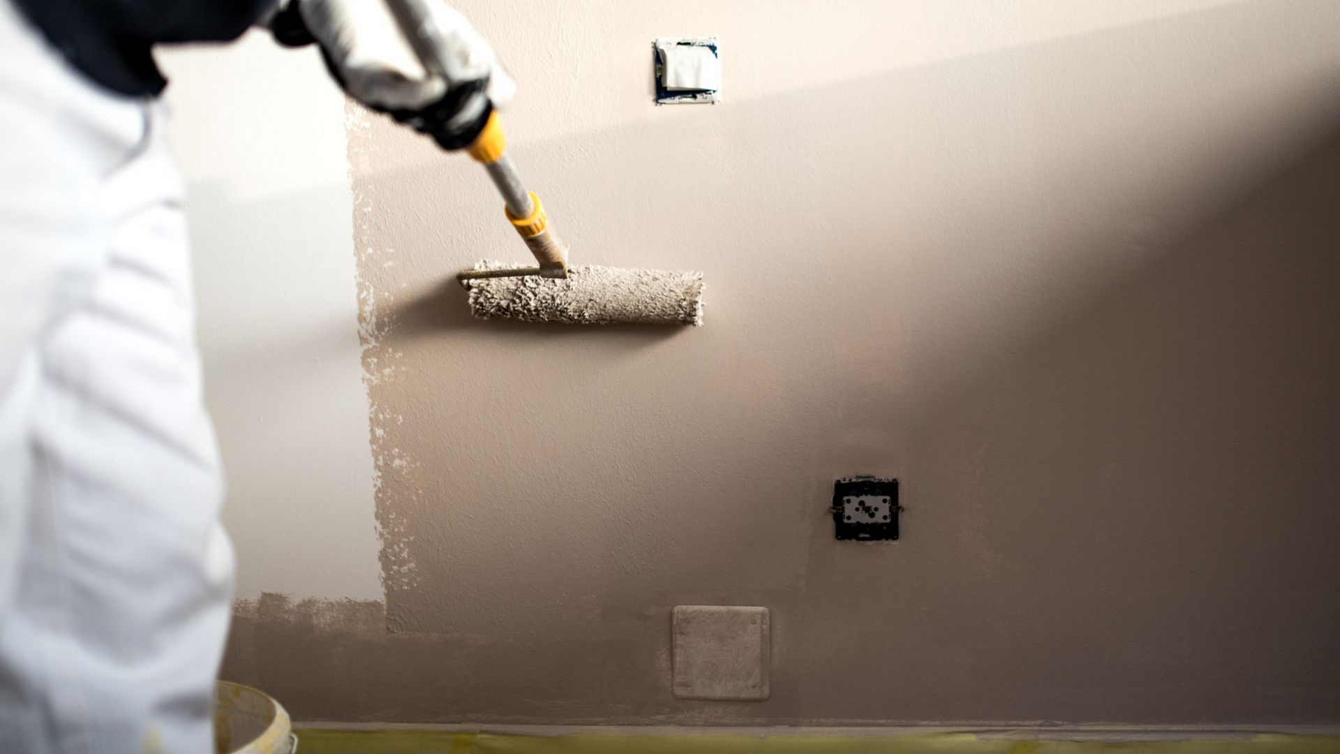 Personne en salopette blanche peignant un mur au rouleau ; couleur de peinture neutre.