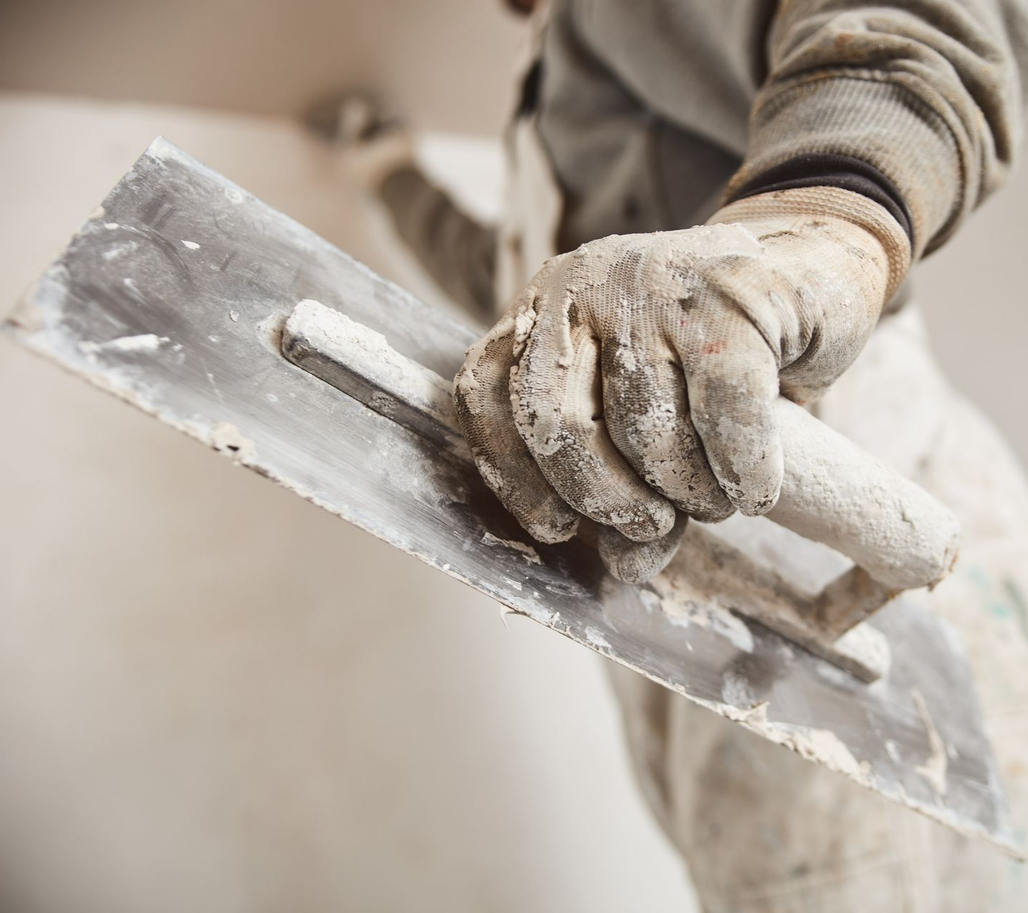 Une main gantée tient un platoir tout en appliquant du plâtre sur un mur.