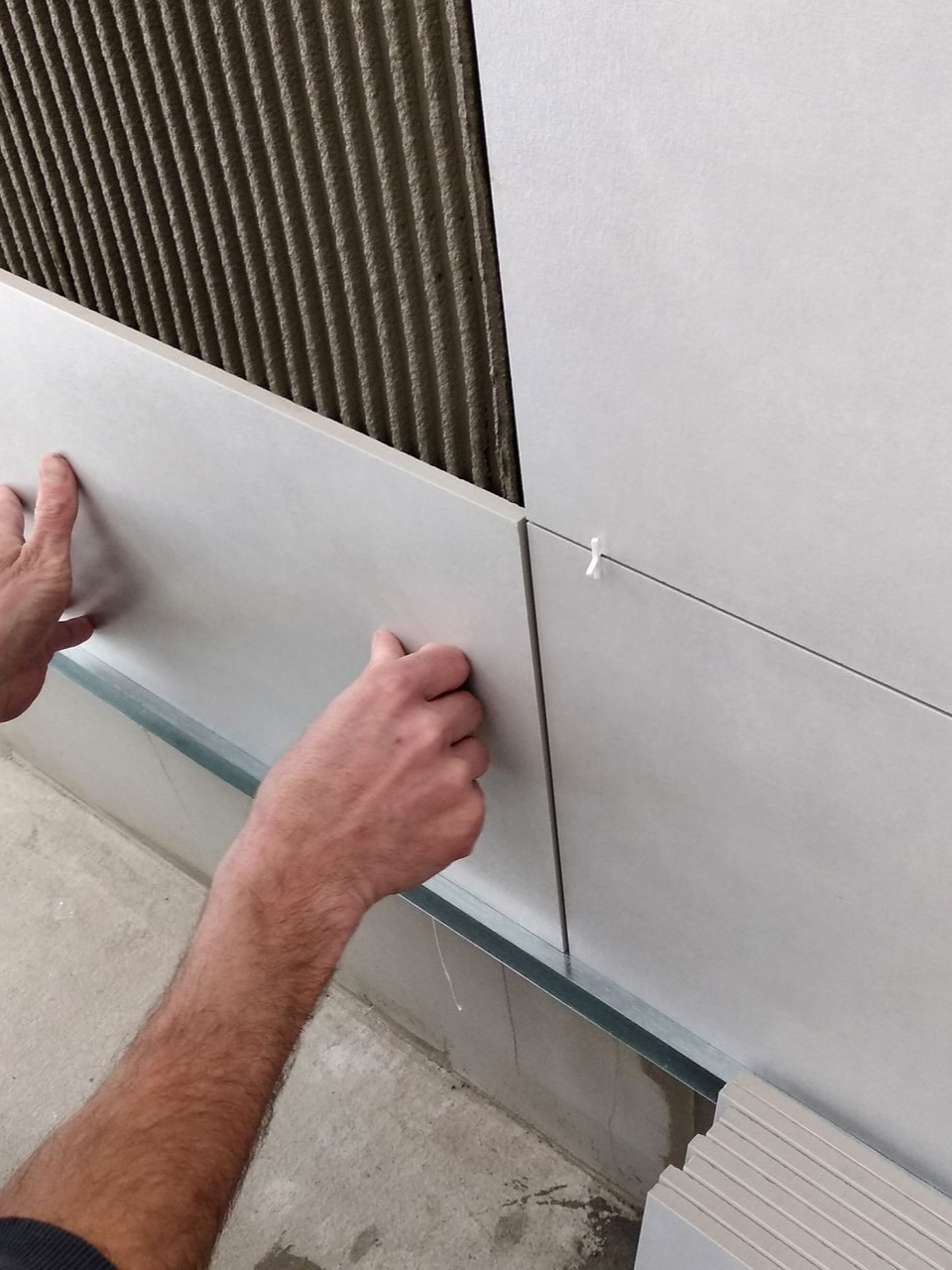 Une personne installe un carreau clair sur un mur, laissant apparaître une couche de colle.