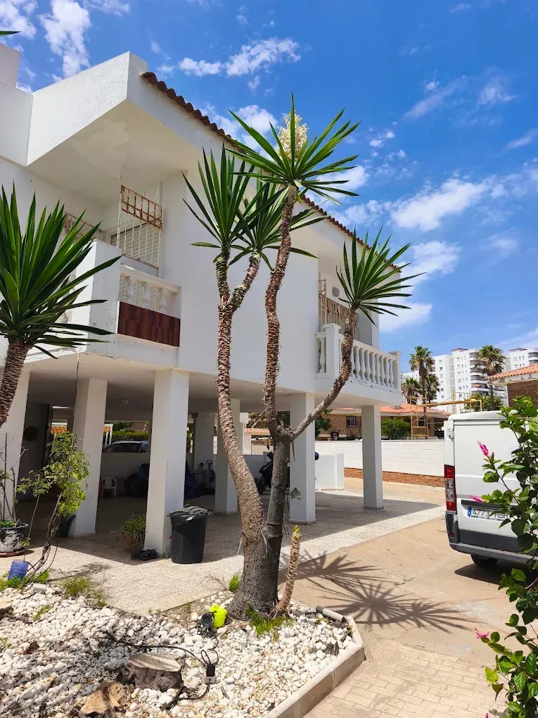 Moderno edificio de apartamentos blanco con palmeras, jardinería con grava y una furgoneta blanca aparcada bajo un cielo azul