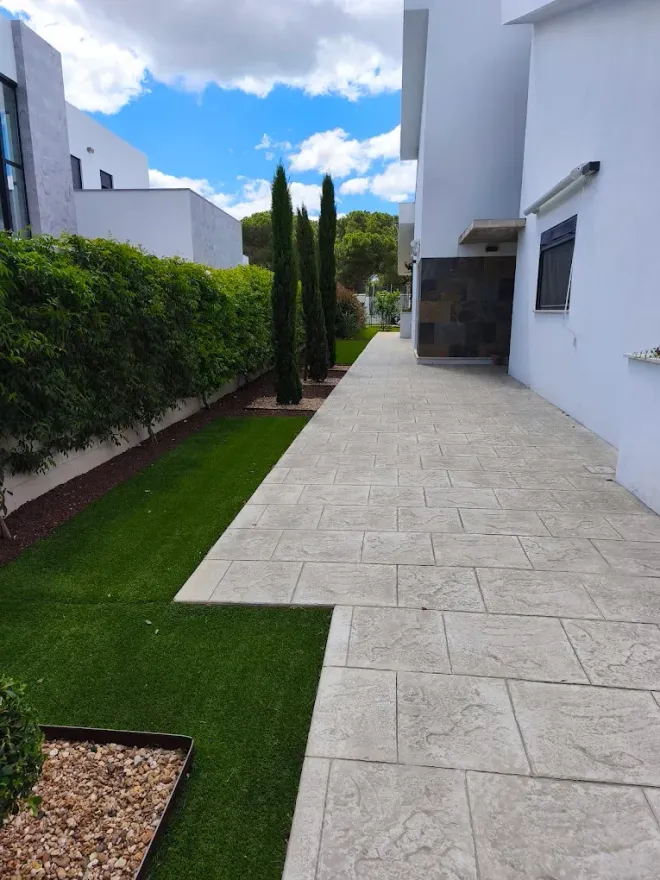 Sendero de una casa moderna con pavimento de piedra blanca, césped verde, setos y altos cipreses bajo un cielo azul.