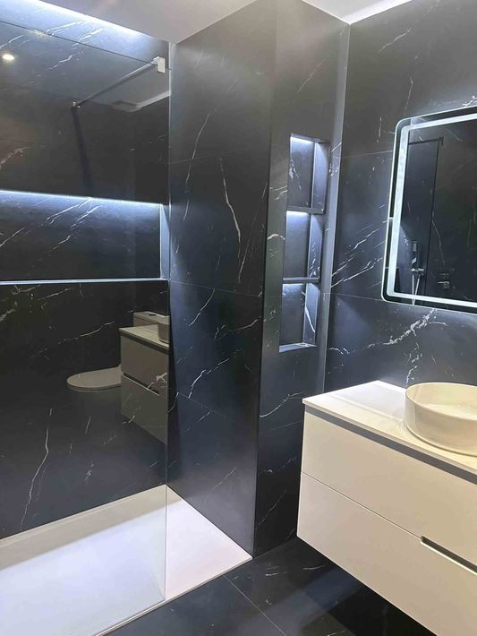 Baño de mármol blanco y negro con lavabo y ducha blancos, estantes iluminados y un espejo con luz de borde.