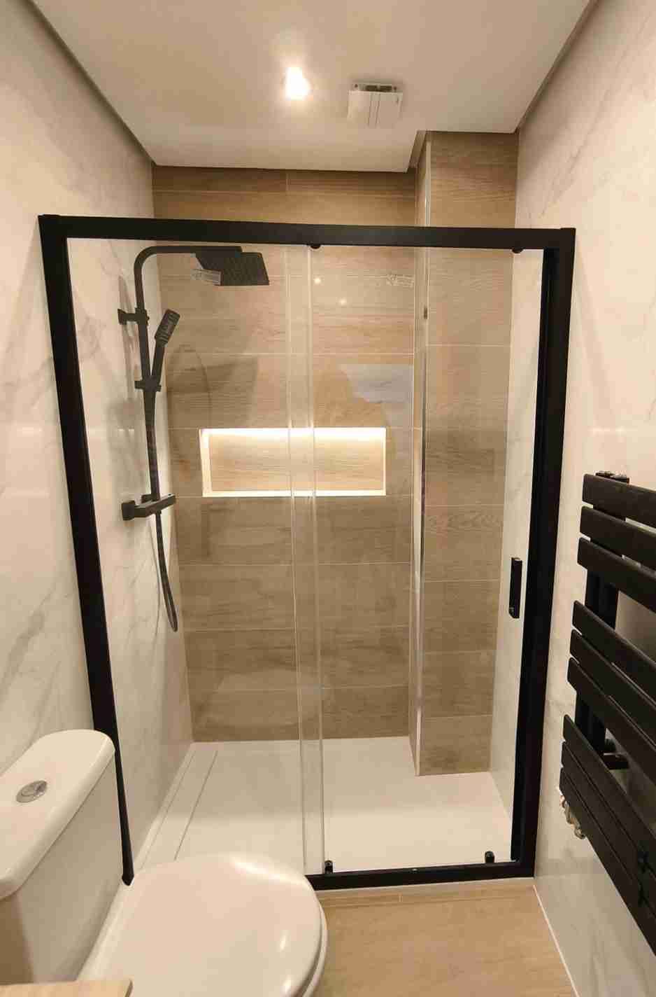 Pequeño baño moderno con ducha de cristal con marco negro, inodoro y toallero negro.
