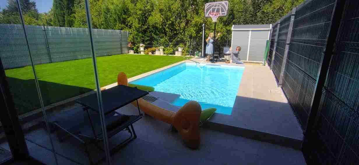 Una piscina en el patio trasero con canasta de baloncesto y césped. Un juguete inflable flota en la piscina.