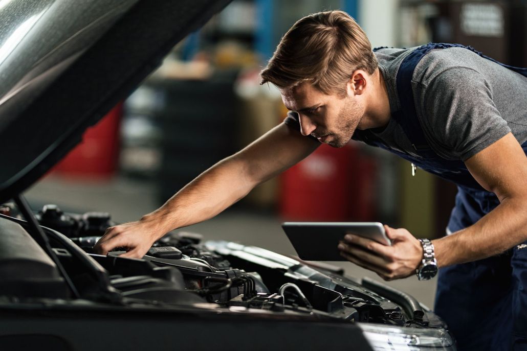 Um mecânico de macacão azul inspeciona o motor de um carro aberto enquanto segura um tablet em uma garagem.