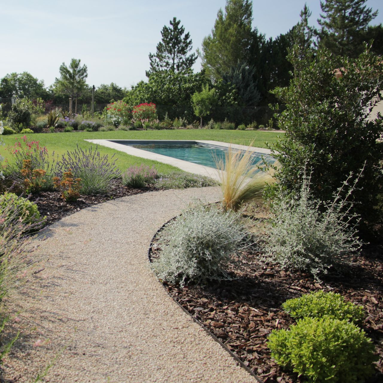 Un chemin de gravier sinueux traverse un jardin planté d'arbustes et recouvert de paillis, menant à une piscine.