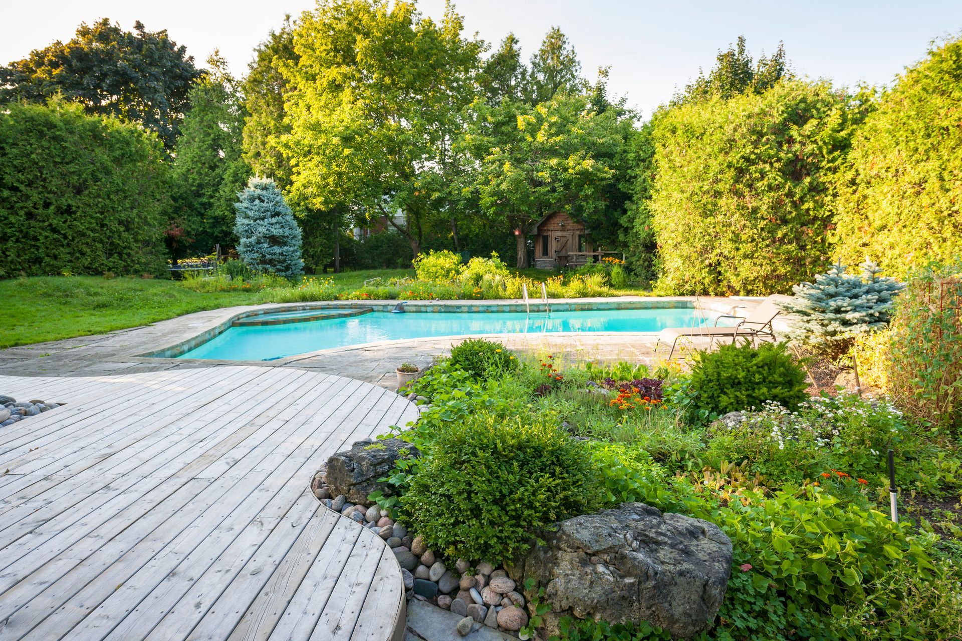 Une terrasse en bois clair devant une piscine turquoise entourée d'arbres verdoyants et de parterres de fleurs.