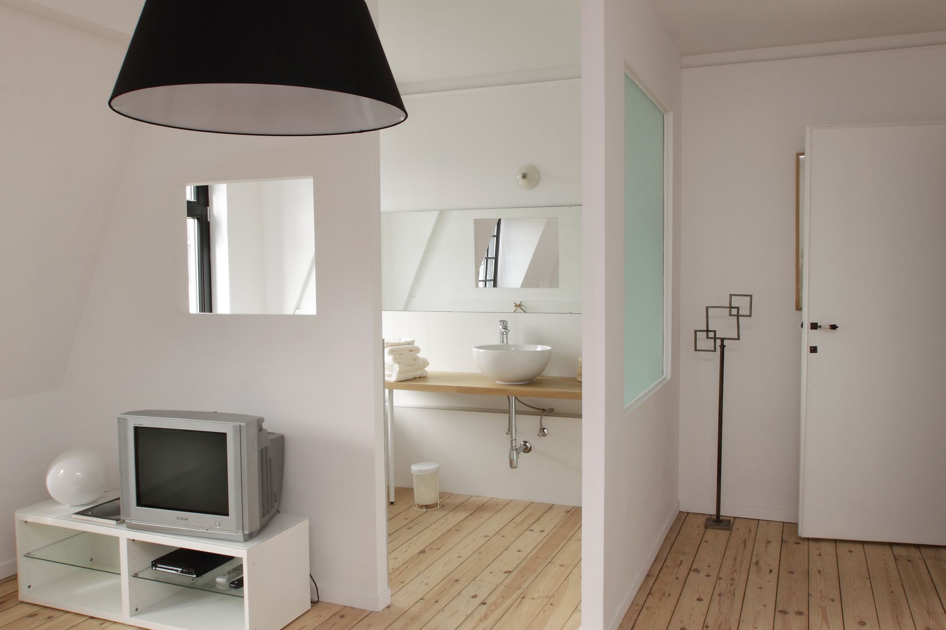 Pièce blanche minimaliste avec parquet, meuble TV, miroir et porte ouverte donnant sur un petit coin lavabo.