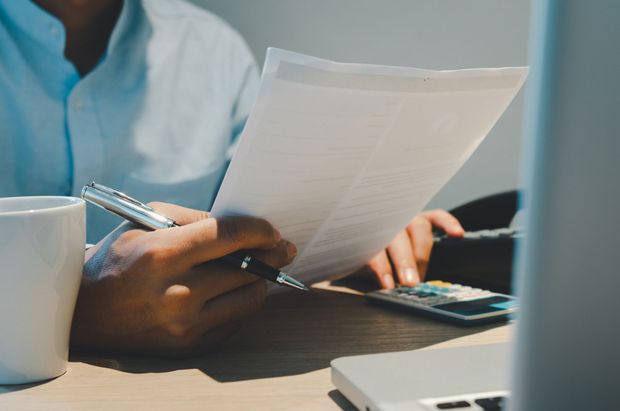 Persona con camisa azul claro revisa documentos, calculando en una calculadora, con una taza de café y una computadora portátil en el escritorio.