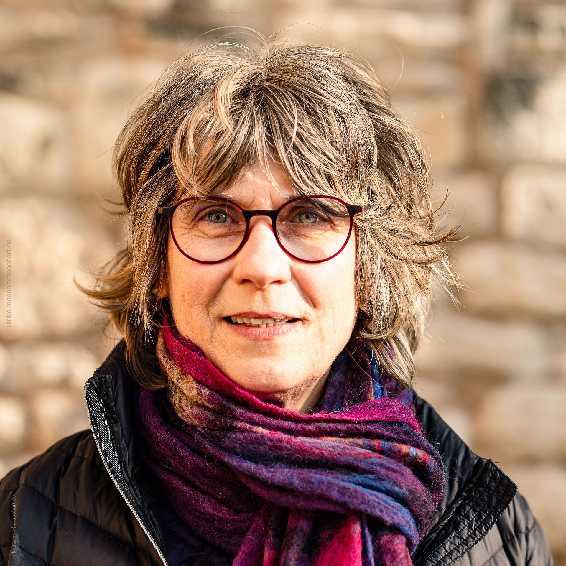 Eine Frau mit Brille und Kopftuch steht vor einer Steinmauer.