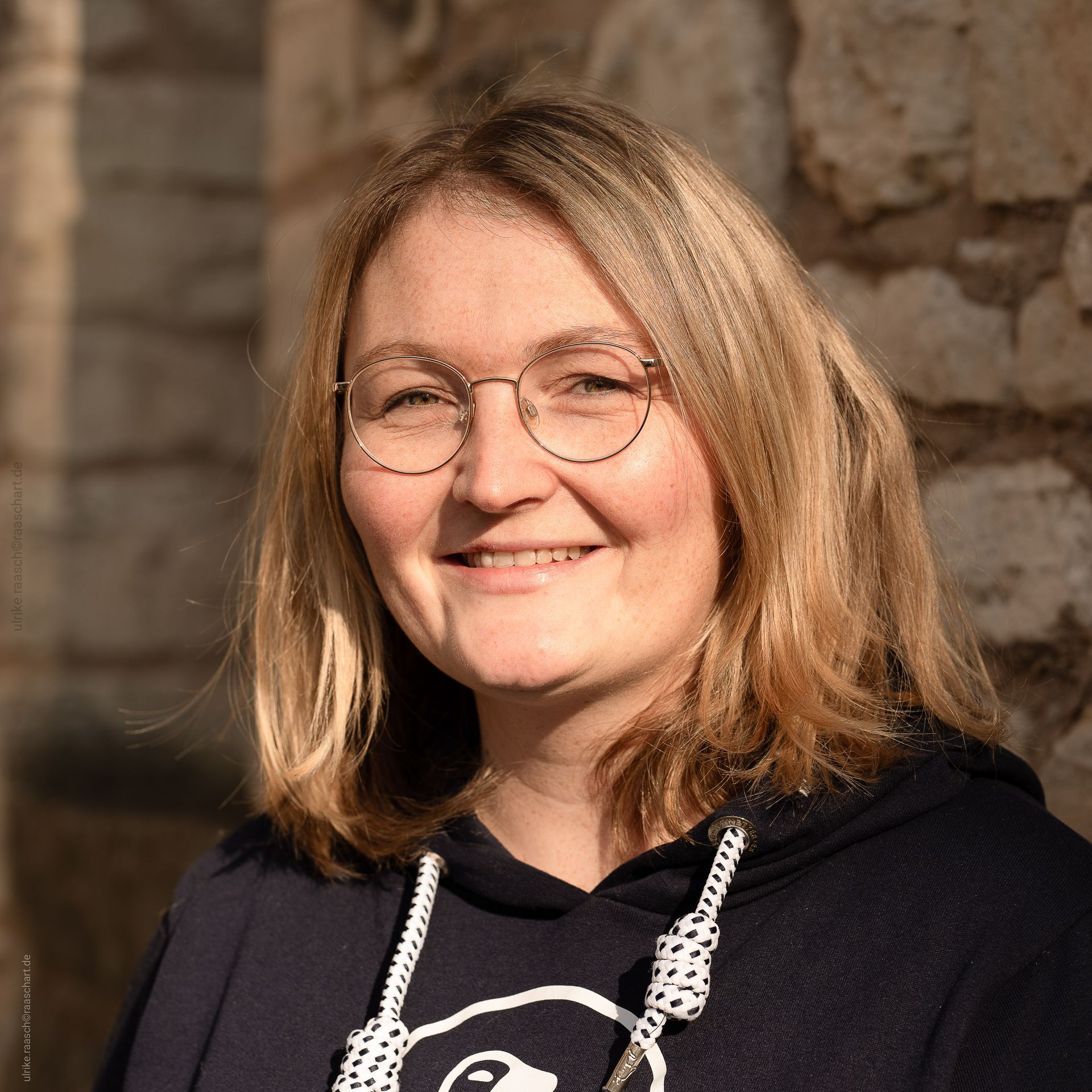 Eine Frau mit Brille und Kapuzenpullover lächelt in die Kamera.