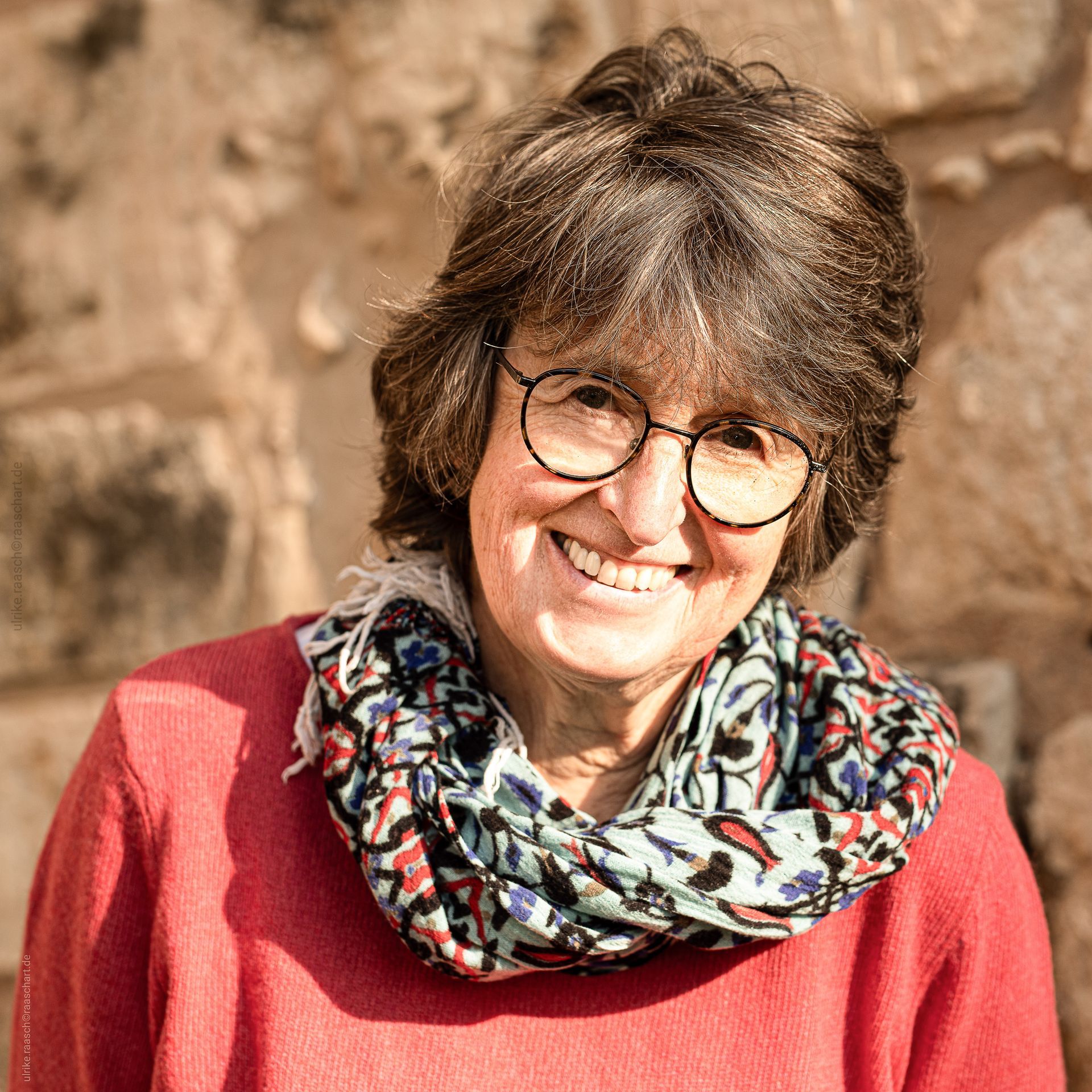Eine Frau mit Brille und Schal lächelt vor einer Steinmauer.