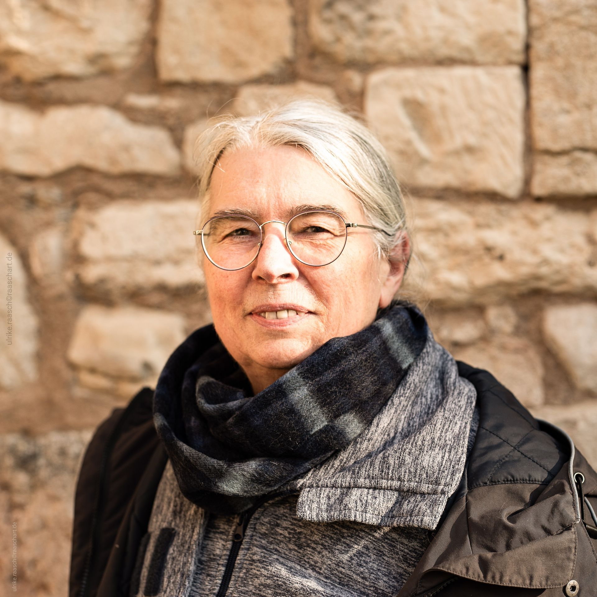 Eine Frau mit Brille und Kopftuch steht vor einer Steinmauer.