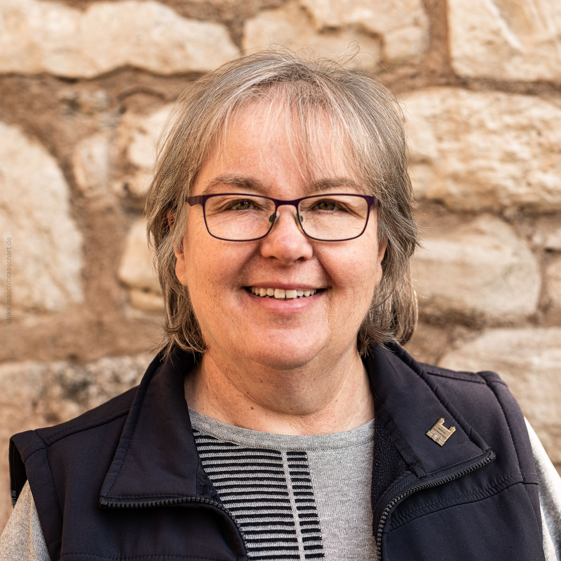 Eine Frau mit Brille und Weste lächelt vor einer Steinmauer.