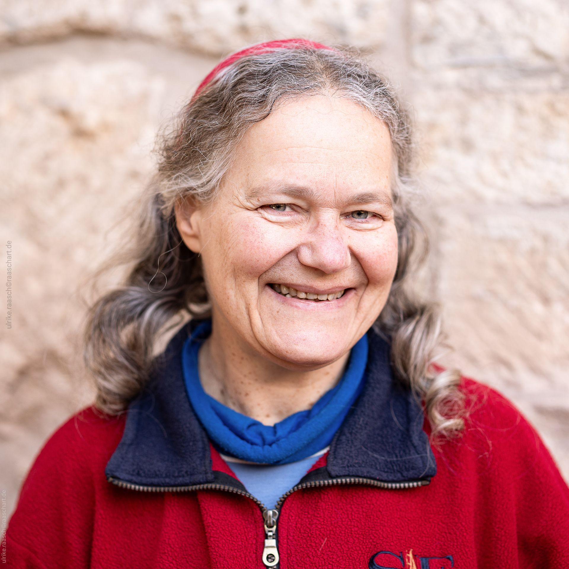 Eine Frau mit einer roten Jacke und einem blauen Schal lächelt.