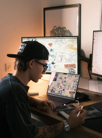Persona con gorra y gafas, trabajando en un escritorio con varios monitores, una computadora portátil y una tableta de dibujo