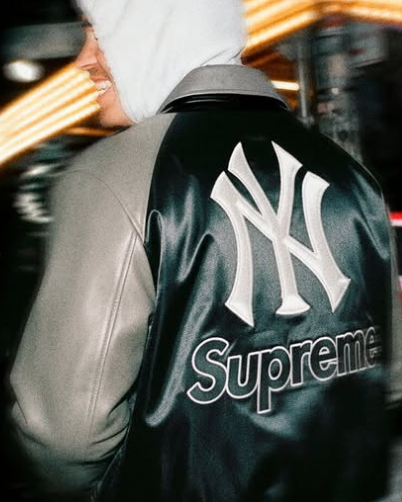 Persona que viste una chaqueta Supreme Yankees negra y gris, al aire libre.