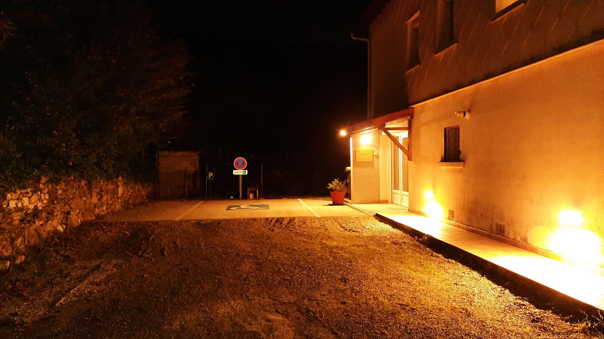 La façade d'un bâtiment faiblement éclairée la nuit. On distingue une bordure, un trottoir et un panneau de signalisation.
