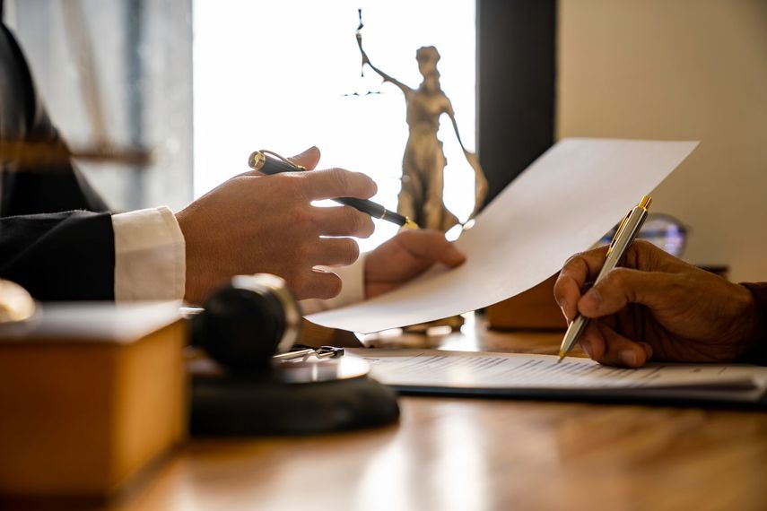 Un avocat examine un document avec son client et signe un contrat. Une statue de la justice apparaît en arrière-plan.