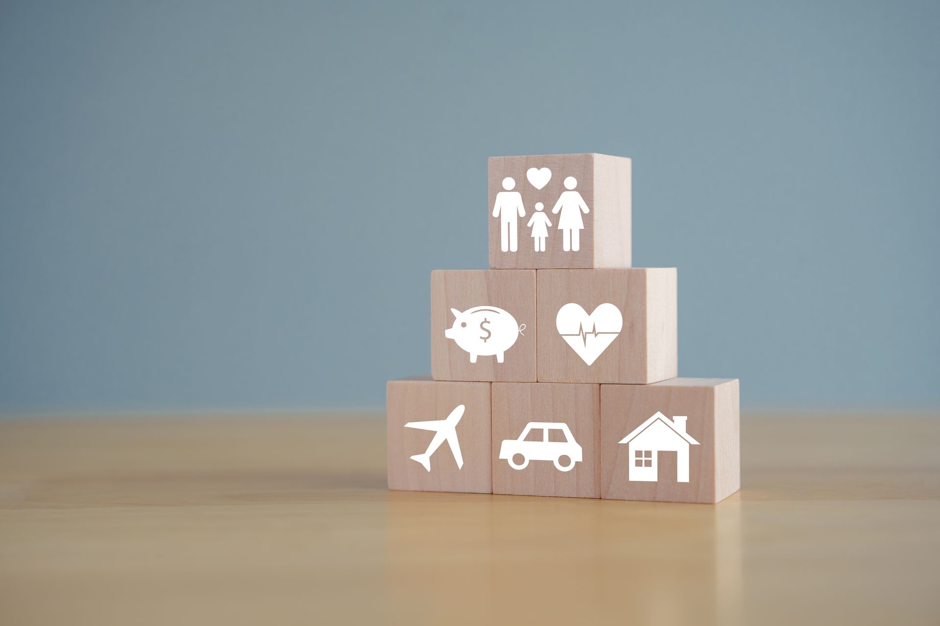 Blocs en bois avec des icônes d'assurance disposées en pyramide : famille, cœur, maison, voiture, avion et tirelire.