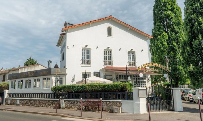 Bâtiment en stuc blanc avec toit de tuiles rouges et grands arbres derrière une clôture basse à un coin de rue.