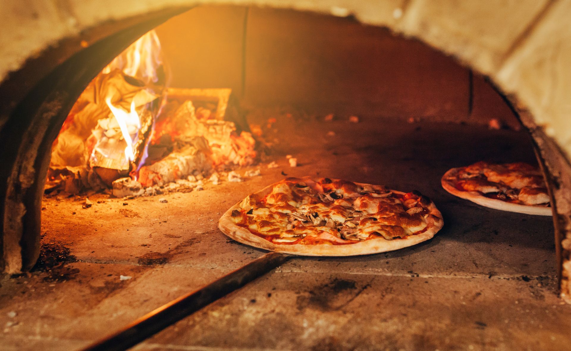 Cuisson de pizzas dans un four à bois, avec des flammes à gauche et deux pizzas sur le sol en pierre.