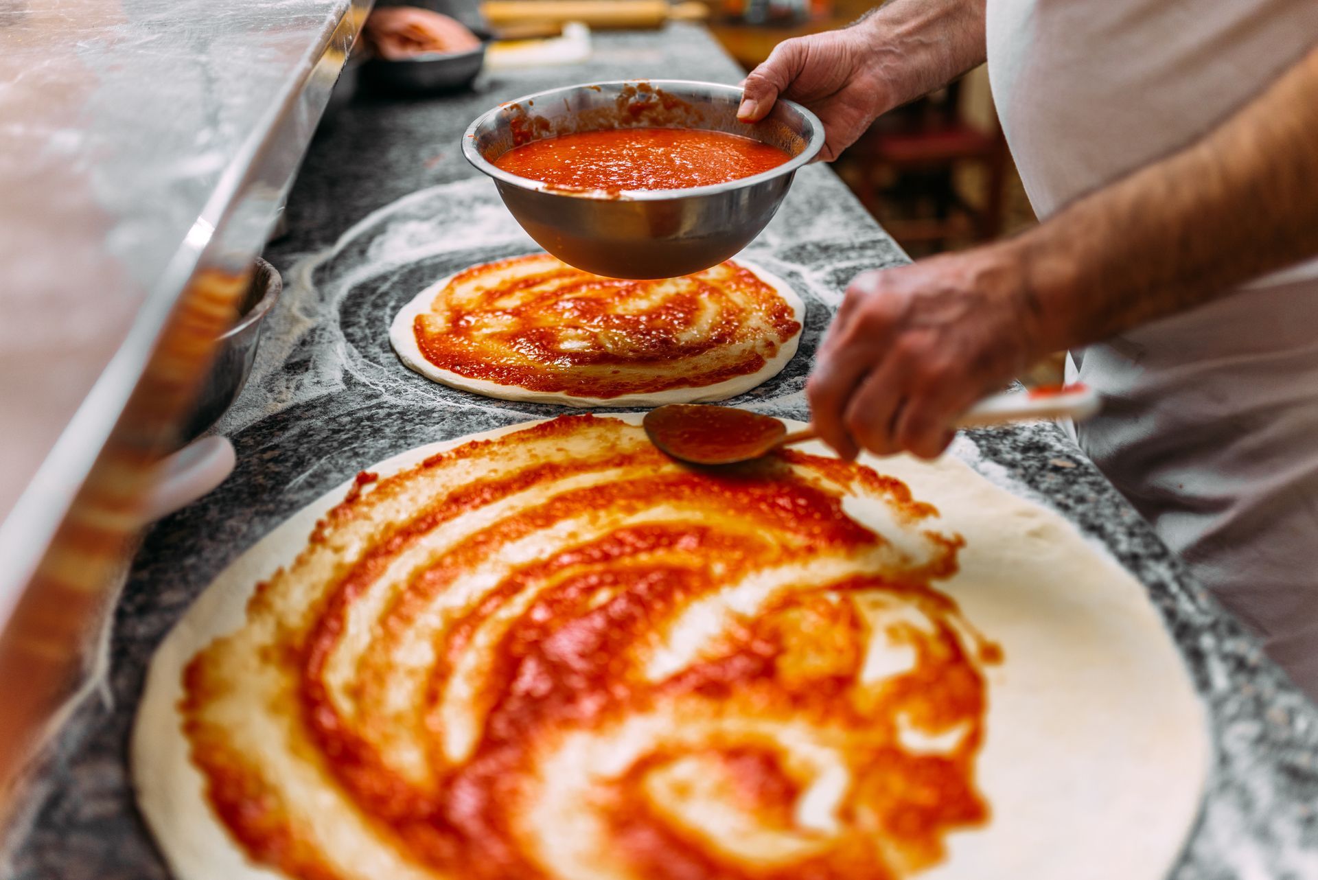 Des mains étalant de la sauce tomate sur une pâte à pizza posée sur un plan de travail fariné dans une cuisine.