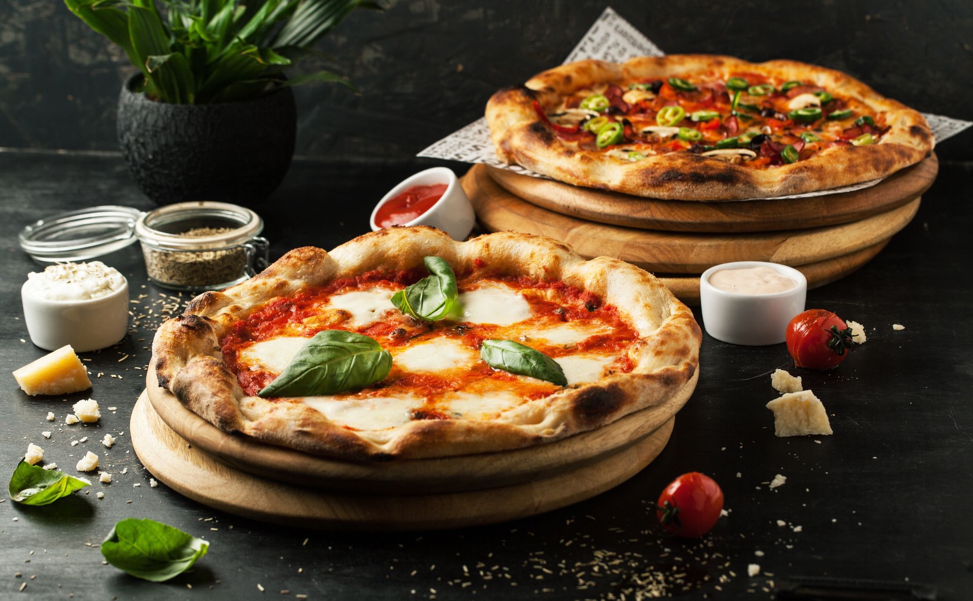 Deux pizzas rustiques sur des planches en bois, garnies de basilic et de tomates, sur une table sombre.