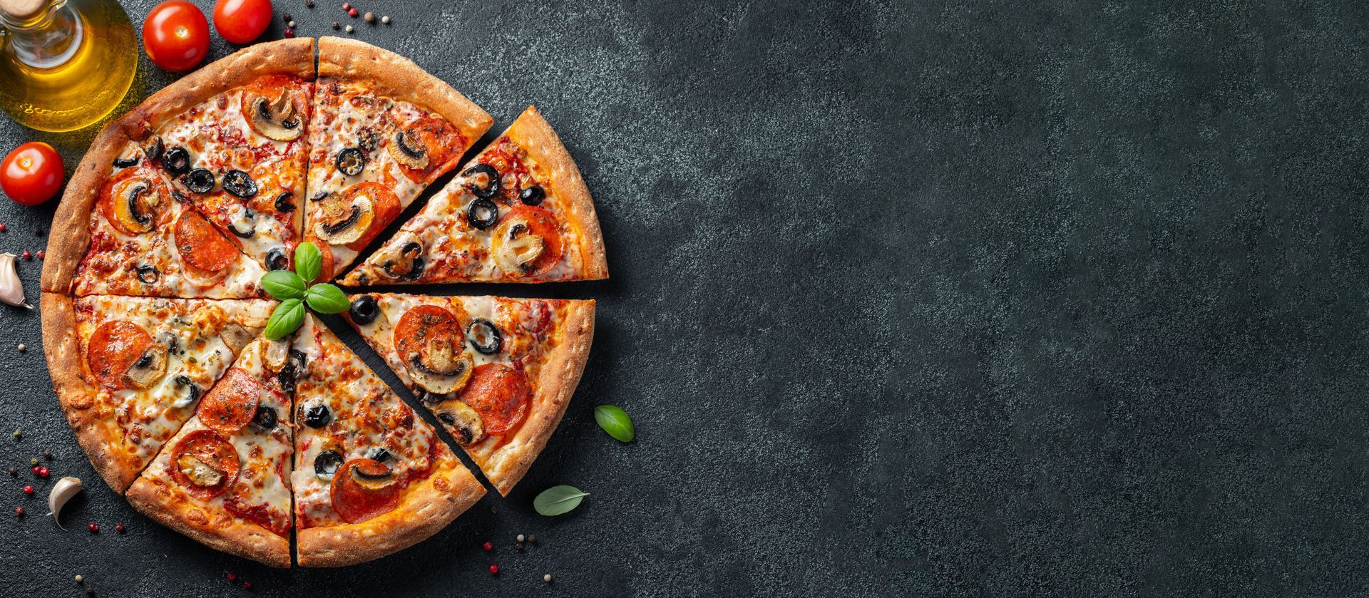 Pizza au fromage coupée en tranches sur une table sombre, garnie de tomates et de feuilles de basilic.