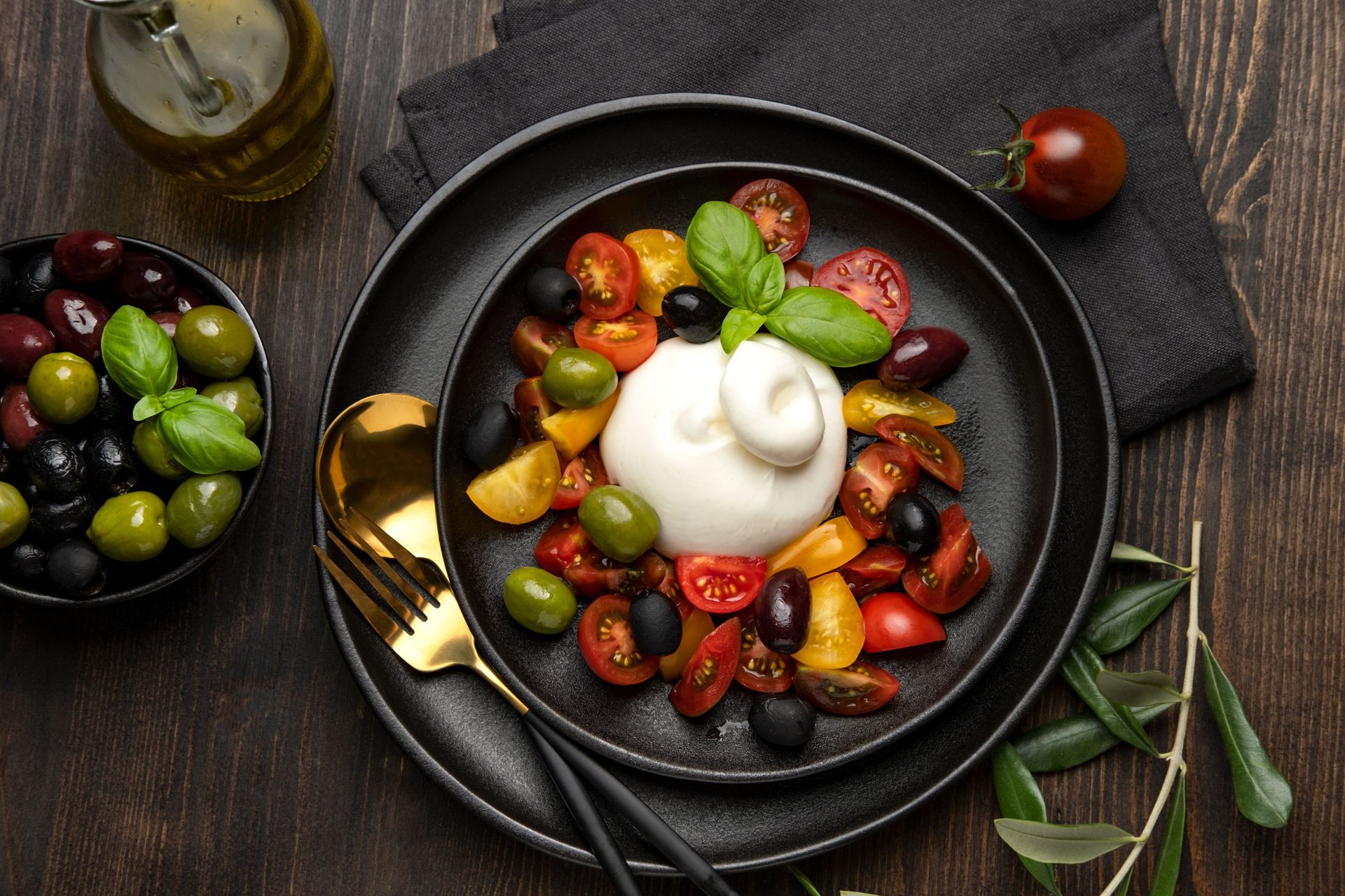 Bol de burrata avec tomates cerises, olives, basilic et huile d'olive sur une table sombre.