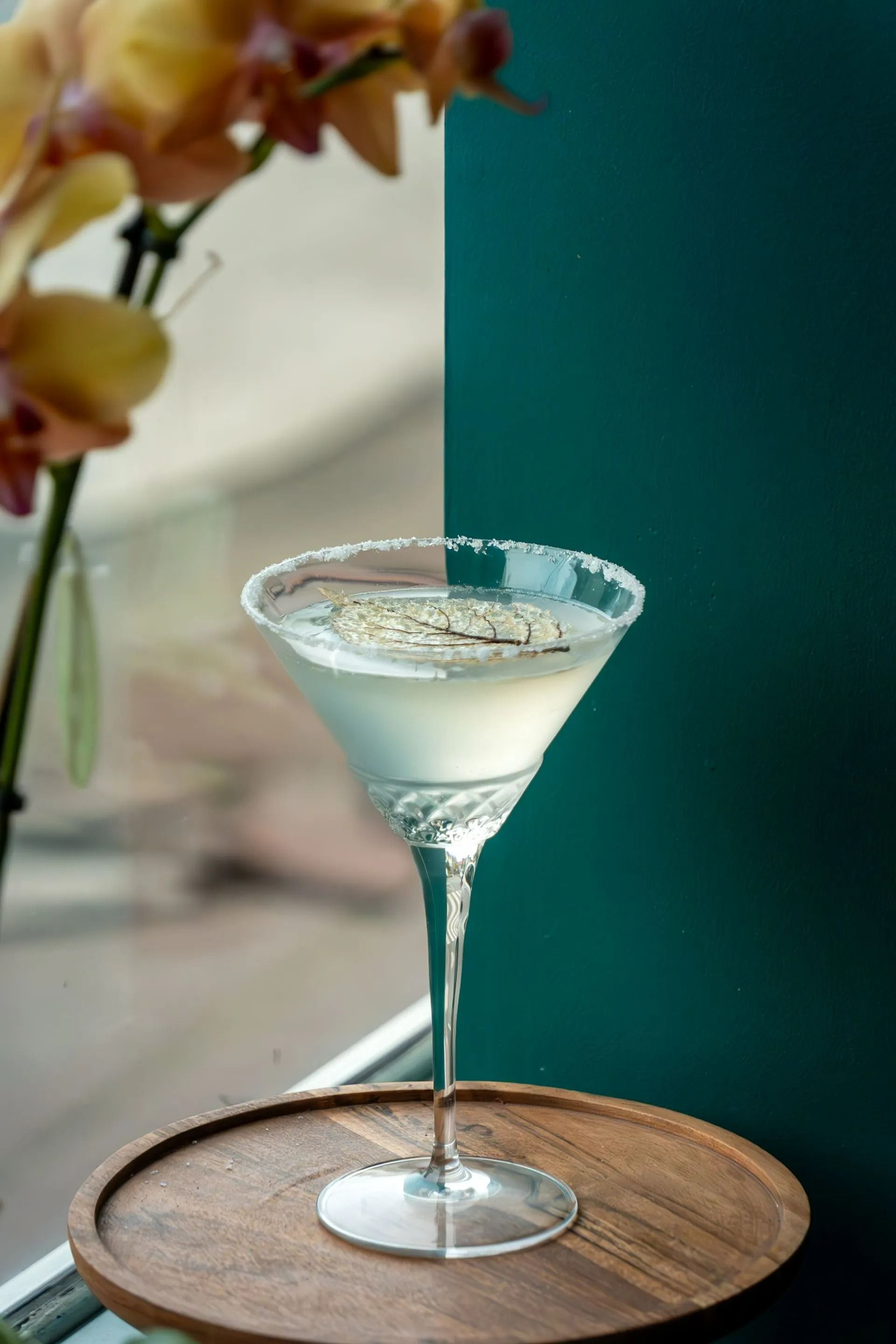 Un verre à cocktail à bord dépoli posé sur un plateau en bois, à côté de fleurs et d'un mur bleu canard foncé.