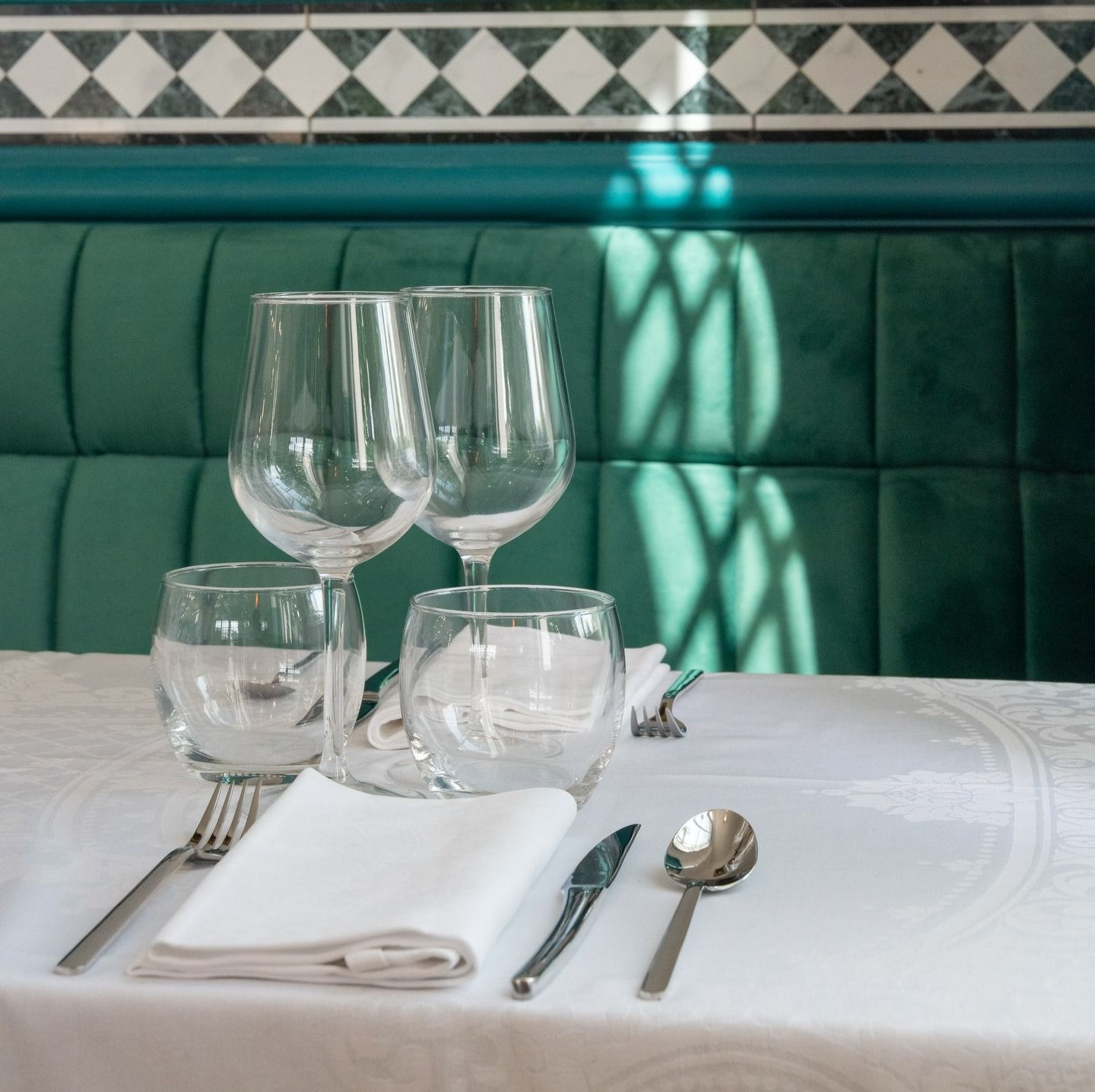 Élégante table dressée avec nappes blanches, verres à vin et couverts en argent dans une banquette verte près d'un mur carrelé.