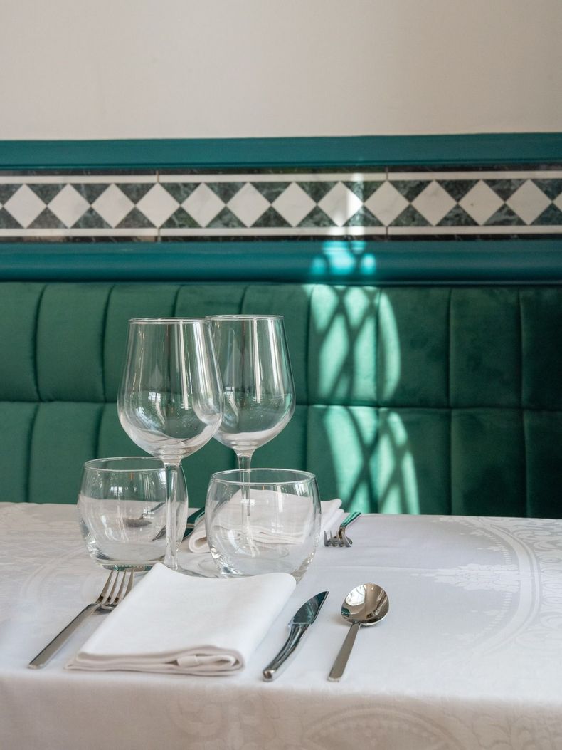 Table dressée avec verres à vin et couverts sur une nappe blanche devant une banquette verte, la lumière du soleil projetant des ombres.