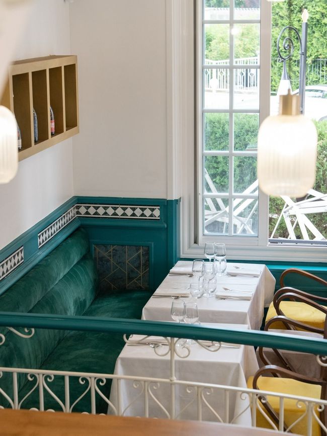 Cabine de café lumineuse avec sièges turquoise, table blanche, chaises jaunes et vue sur la verdure par la fenêtre.