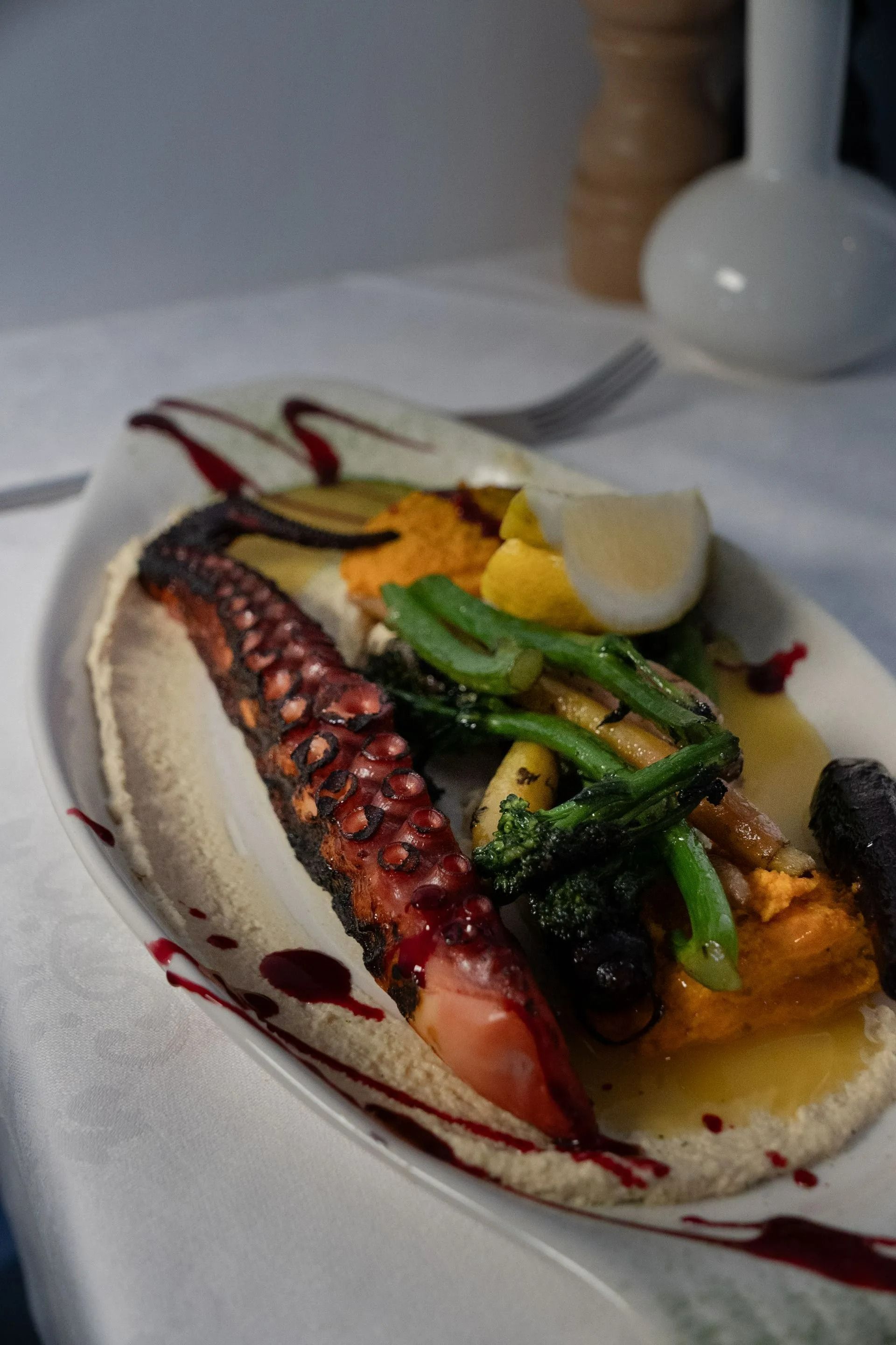 Assiette de fruits de mer avec tentacule de poulpe, légumes verts, sauce et quartier de citron sur une table blanche.
